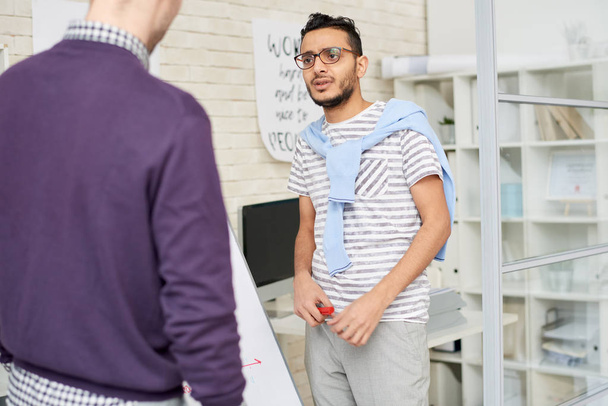 Portraitaufnahme eines gut aussehenden Mixed-Race-Designers in Casualwear, der kreative Ideen mit einem Kollegen teilt, während er an der Markierungstafel steht, Büroeinrichtung im Hintergrund - Foto, Bild
