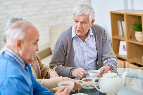 Portrait of white-haired senior man talking to friend while drinking tea at table in modern retirement home, copy space - Photo, image