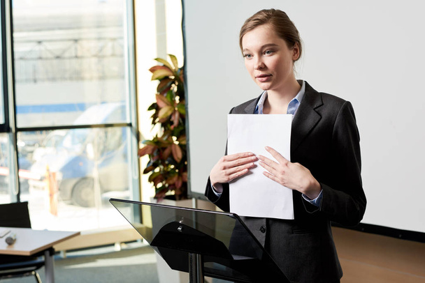 Waist up portrait of young businesswoman standing at podium giving presentation to audience - Foto, afbeelding