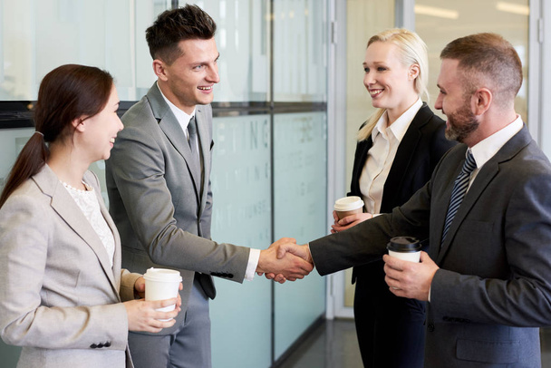 Retrato de cuatro socios de negocios estrechando la mano y sonriendo de pie en el vestíbulo del edificio de oficinas moderno después de cerrar acuerdo beneficioso
 - Foto, imagen