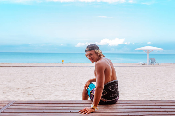 Le surfeur est assis sur la terrasse de la plage dos à la caméra et tourne le visage en regardant la caméra
 - Photo, image