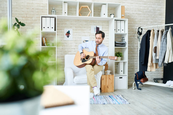 Stylowy mężczyzna siedzi w creative studio modnej i gra na gitarze na biały wygodny fotel. - Zdjęcie, obraz