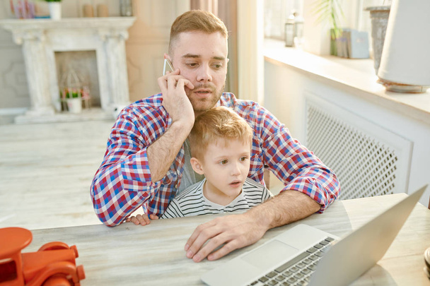 Warm toned portrait of handsome modern man trying to work while caring for his son at home - Photo, Image