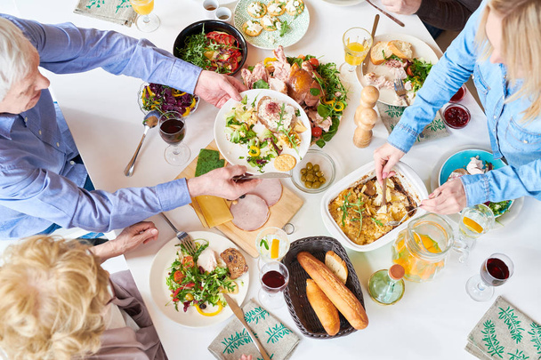 Вид на большую счастливую семью, сидящую за обеденным столом и наслаждающуюся вкусной домашней едой во время праздничного торжества и раздачу тарелок за столом
 - Фото, изображение