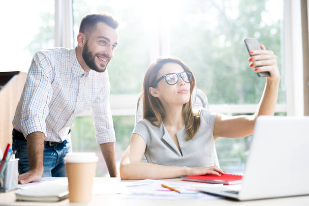 Portrait of beautiful businesswoman wearing glasses taking selfie  with handsome colleague while sitting at desk in office  against window in sunlight - Photo, Image