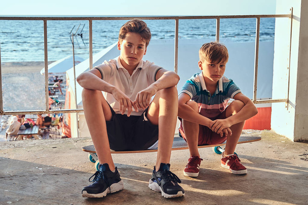 Due fratellini seduti insieme su uno skateboard vicino a guardrail sullo sfondo della costa
. - Foto, immagini