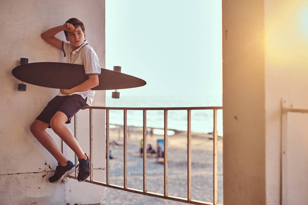 Ritratto di un giovane skater boy che tiene uno skateboard seduto su un guardrail sullo sfondo di una spiaggia al tramonto
. - Foto, immagini