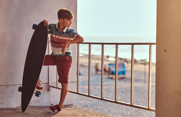 Portret van een jonge skater jongen gekleed in t-shirt en short leunend op de vangrail en bezit een skateboard tegen de achtergrond van een Zeekust bij de zonsondergang. - Foto, afbeelding