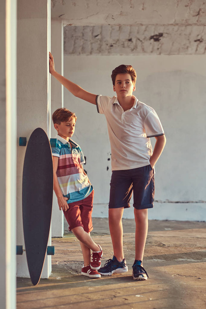 Portret van twee kleine broers met een skateboard leunend op een muur buiten bij een zonsondergang. - Foto, afbeelding