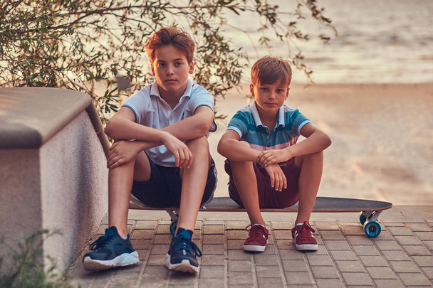 Портрет двух маленьких братьев, сидящих вместе на скейтборде на фоне морского побережья на закате
. - Фото, изображение