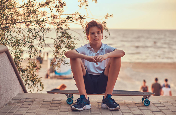 Fiatal deszkás srác ül egy gördeszka, a háttérben egy tengerpart naplemente. - Fotó, kép