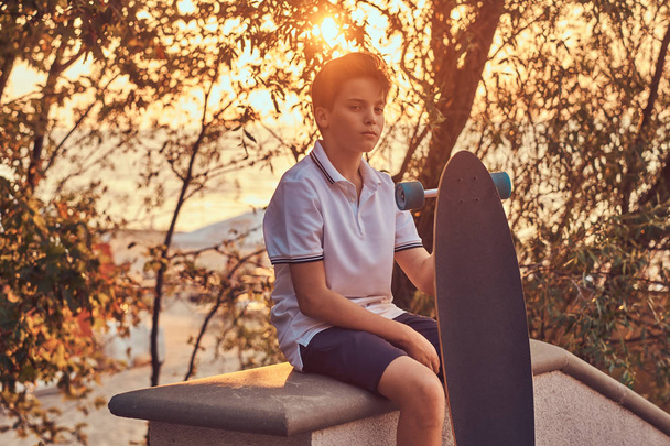 Junge Skater in T-Shirt und kurzer Hose sitzt bei Sonnenuntergang auf der steinernen Leitplanke.  - Foto, Bild