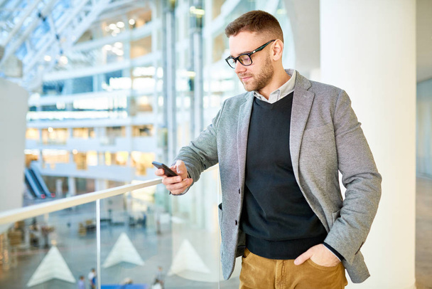 handsome bearded man wearing stylish clothes texting with friend on smartphone while standing on second floor of modern shopping mall - Foto, imagen