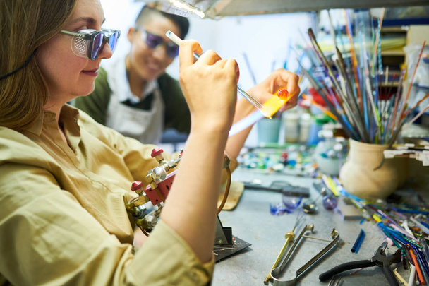 Портрет молодой художницы, которая плавит стекло газовой горелкой, делая бусины для красивых ювелирных изделий ручной работы в стекольной мастерской
 - Фото, изображение