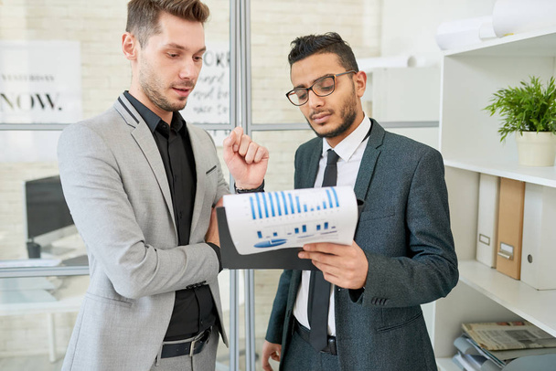 Handsome mixed-race white collar worker showing statistic data to male colleague while having working meeting at open plan office - Photo, Image