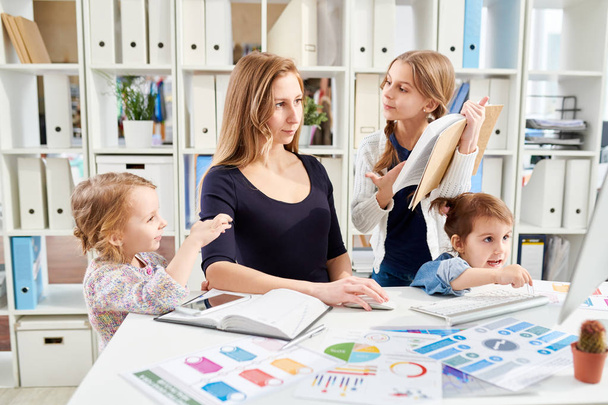 Young white collar worker sitting at desk and creating visual presentation for business partners while her little daughters trying to attract her attention, office interior on background - Фото, зображення