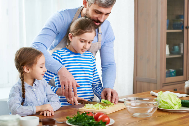 Προετοιμάζετε το δείπνο στην ευρύχωρη κουζίνα: όμορφο κορίτσι στέκεται στο ξύλινο τραπέζι και κοπή λάχανο ενώ τον γενειοφόρος μεσήλικας μπαμπά της, βοηθώντας την μικρή αδελφή που παρακολουθεί τους - Φωτογραφία, εικόνα