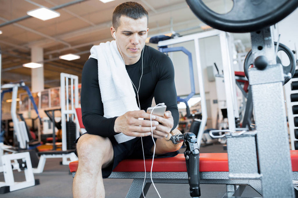 Портрет красивого мускулистого мужчины с протезной ногой, сидящего на скамейке и расслабляющего слушая музыку со смартфона после тренировки в современном тренажерном зале
 - Фото, изображение