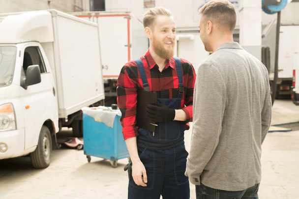 Young bearded male auto technician in workwear standing in repair shop, talking to his acquaintance and smiling happily - Photo, Image