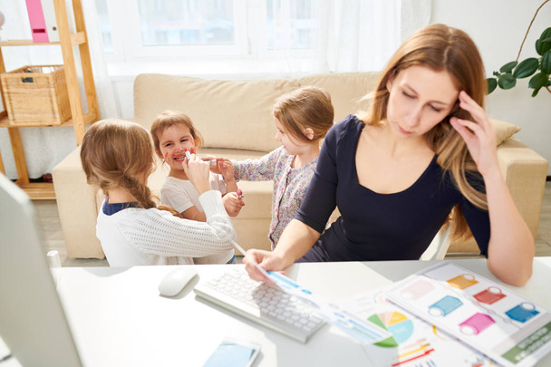 Busy manager sitting at desk and analyzing statistic data while her little daughters coloring their faces with red lipstick, interior of cozy living room on background - Foto, Bild
