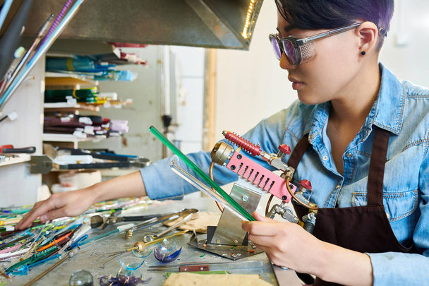 Seitenansicht einer asiatischen jungen Frau, die in einer modernen Werkstatt nach Werkzeugen greift, während sie mit Glas arbeitet, Kopierraum - Foto, Bild