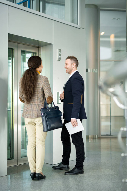 Täyspitkä muotokuva kahdesta liikemiehestä, mies ja nainen juttelemassa iloisesti odottaessaan hissiä toimistorakennuksessa
 - Valokuva, kuva