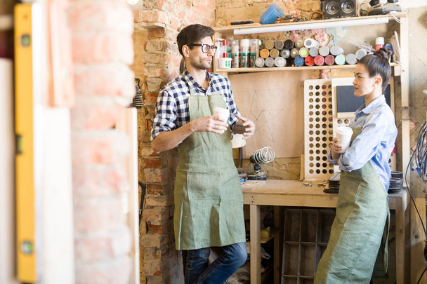 Retrato de dos artesanos modernos que usan delantales hombre y mujer charlando durante el descanso del café mientras trabajan en el taller de elaboración
 - Foto, Imagen