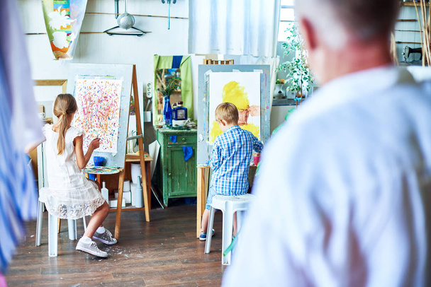 Visão traseira retrato de professor sênior assistindo crianças pintando em cavaletes durante a aula de arte em estúdio acolhedor decorado com plantas
 - Foto, Imagem