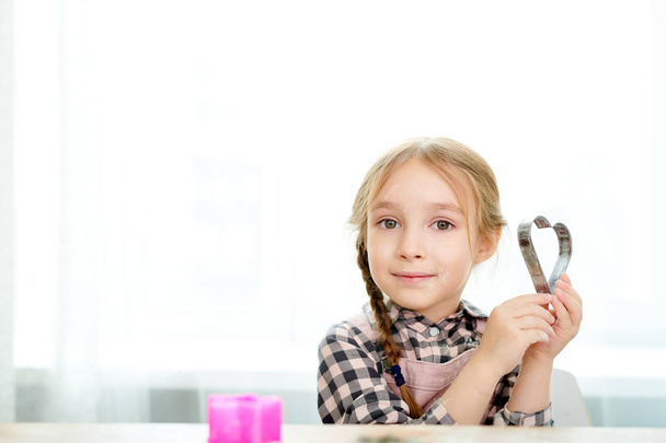 Retrato de linda niña sosteniendo cortador de galletas en forma de corazón y mirando a la cámara mientras disfruta de la cocina en casa, espacio de copia
 - Foto, imagen