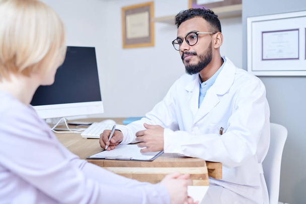 Retrato del guapo médico de Oriente Medio consultando a una paciente sentada en el escritorio en una oficina moderna, espacio para copiar
 - Foto, imagen