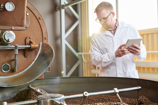 Retrato de cintura hacia arriba del trabajador moderno que usa bata de laboratorio que trabaja en la fábrica de producción de café, escena iluminada por la luz del sol
 - Foto, imagen