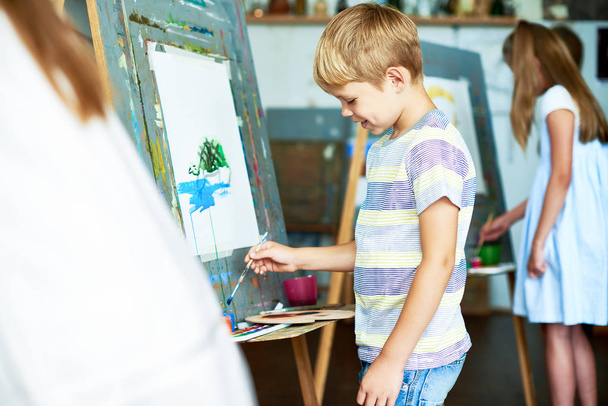 Πλευρά πορτρέτο θέα της χαριτωμένο μικρό ξανθό αγόρι ζωγραφική σε καβαλέτο κατά τη διάρκεια της κατηγορίας τέχνης και χαμογελώντας ευτυχώς - Φωτογραφία, εικόνα