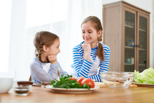 lächelndes kleines Mädchen mit Schürzen, die sich anschauen, während sie das festliche Menü für die Muttertagsfeier besprechen, Innenraum der geräumigen Küche im Hintergrund - Foto, Bild