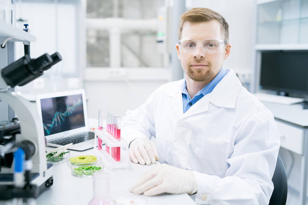 Portret van lachende jonge wetenschapper dragen laboratoriumjas camera kijken tijdens het werken aan onderzoek in laboratorium - Foto, afbeelding
