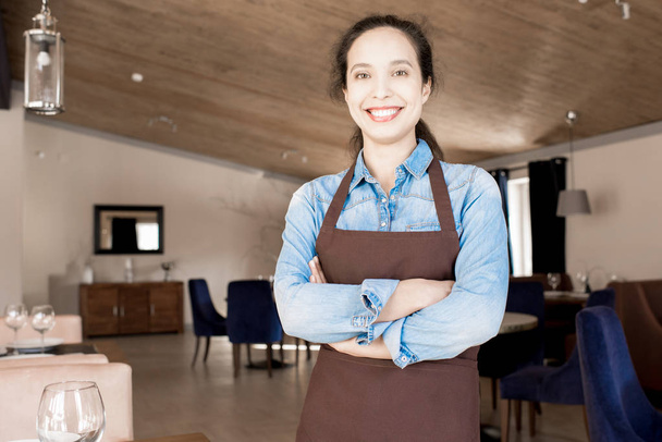 Happy jolly attractive young Hispanic female student wearing apron working as waitress in restaurant, public establishment staff - 写真・画像