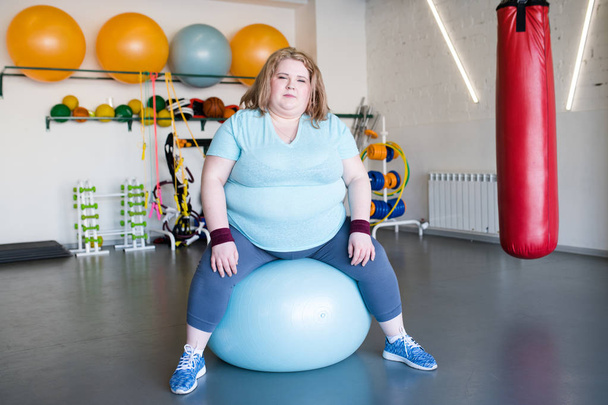 Full length portrait of young obese woman sitting on fitness ball looking at camera - Фото, зображення