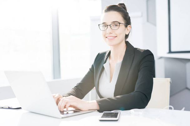 Portret van succesvolle jonge zakenvrouw met behulp van de laptop terwijl het werken bij de balie in kantoor, camera kijken en glimlachen - Foto, afbeelding