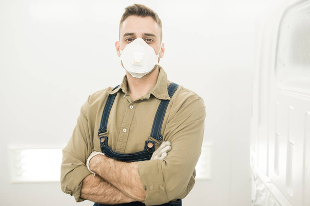 Retrato de jovem técnico de automóveis do sexo masculino em máscara protetora em pé na oficina de reparação com as mãos cruzadas e olhando para a câmera
 - Foto, Imagem