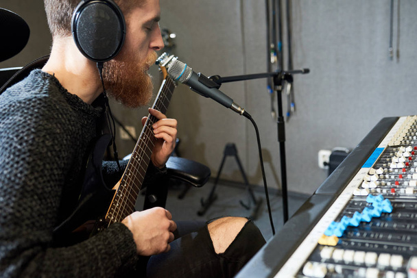 Widok z boku człowieka w słuchawkach gra na gitarze i śpiewa do mikrofonu, siedząc w konsoli w studio nagrań.  - Zdjęcie, obraz