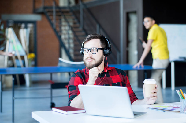 Retrato de homem barbudo moderno usando óculos e camisa vermelha usando laptop olhando para longe pensivamente no escritório de espaço aberto da equipe de desenvolvedores de TI, espaço de cópia
 - Foto, Imagem