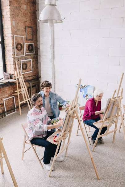High angle portrait of adult students  painting sitting by easels in art studio with female art teacher - Photo, Image