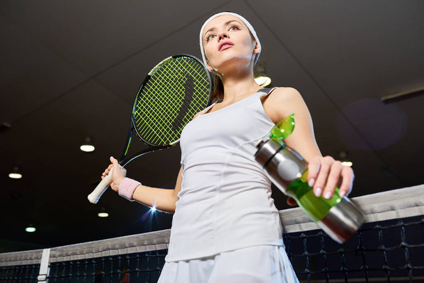 Lage hoek portret van professionele tennisspeelster nemen van onderbreking van de praktijk staande tegen net en drinkwater in overdekte Hof - Foto, afbeelding