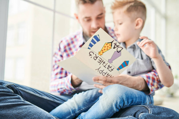 Handsome bearded man slightly embracing his little son while reading Fathers Day greeting card with interest, panoramic window on background - Foto, Imagem