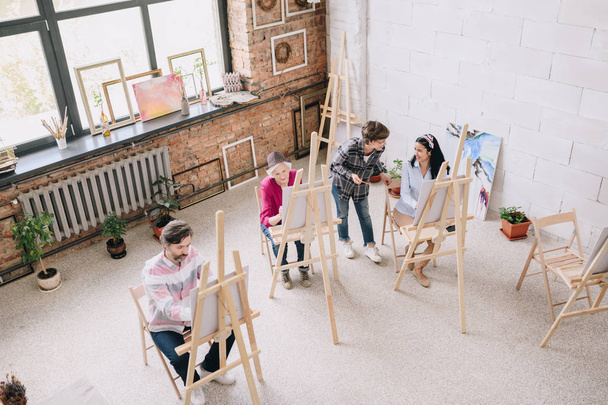 Παραπάνω άποψη πορτραίτο των φοιτητών ζωγραφική κάθεται από καβαλέτα στο art studio με γυναίκα εκπαιδευτικός, βλέποντας τους σε ευρύχωρα, ηλιόλουστα πατάρι χώρου - Φωτογραφία, εικόνα