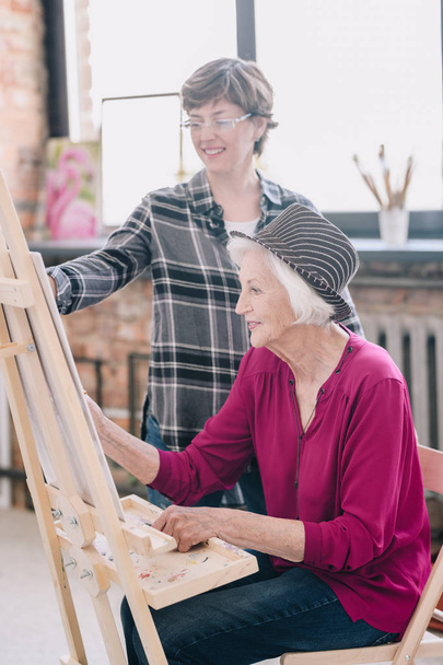 Side view portrait of elegant senior woman painting sitting at easel in art studio and enjoying art with smiling female teacher giving comments, scene in sunlit loft space against big windows. - Photo, Image