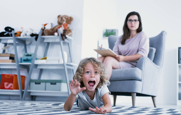 An overactive child screaming and hitting a floor while a psychotherapist is making a diagnose during a meeting in a family support center. - Photo, Image