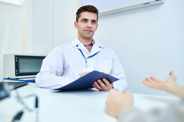 Portrait of young doctor holding clipboard  listening to patients symptoms while sitting at desk in office, copy space - Foto, Imagem
