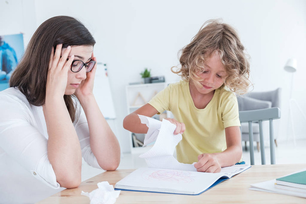 Stressed psychotherapist having a headache during a meeting with a rebellious child with behavioral disorders. The kid is tearing sheets of paper from the notebook. - Foto, Imagen