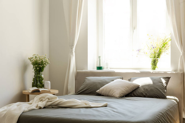 Blanket and pillows on grey bed in bright bedroom interior with flowers and window. Real photo - Фото, изображение