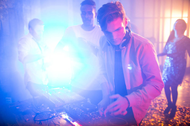 Talentoso jovem DJ usando fones de ouvido misturando música com a ajuda de equipamentos, enquanto envolto em trabalho no clube noturno moderno, grupo de amigos dançando, fundo escuro
 - Foto, Imagem